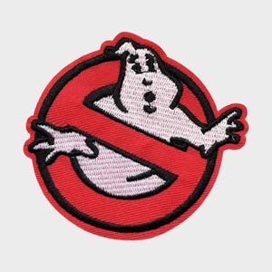 May include: Embroidered patch featuring the Ghostbusters logo. The design includes a white ghost figure inside a red circle with a black border. The ghost has a friendly expression, with arms outstretched.