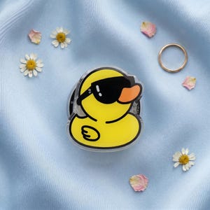 May include: A yellow duck-shaped phone grip with black sunglasses and an orange beak. The phone accessory is on a light blue fabric, surrounded by daisies, rose petals, and a gold ring.