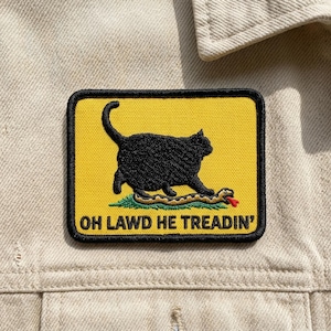 May include: A rectangular patch with a black cat stepping on a snake, set against a yellow background. The patch has a black border and the text "OH LAWD HE TREADIN'" in white.