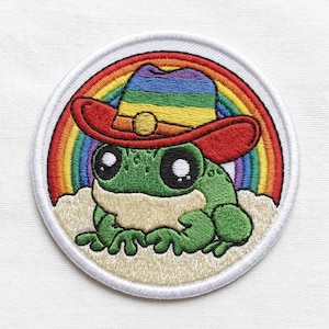 May include: Embroidered patch featuring a green frog wearing a red cowboy hat with a rainbow brim. A rainbow arches behind the frog, and the patch has a white border. The frog is sitting on a beige cloud.