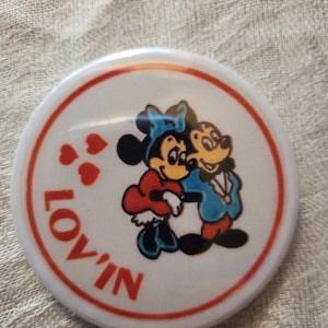 Op de afbeelding: Een ronde witte knoop met een rode rand en het woord "LOV'IN" in het rood. De knoop toont een cartoonafbeelding van Mickey en Minnie Mouse die elkaar omhelzen, met rode harten. De personages dragen blauwe, rode en gele kleding.