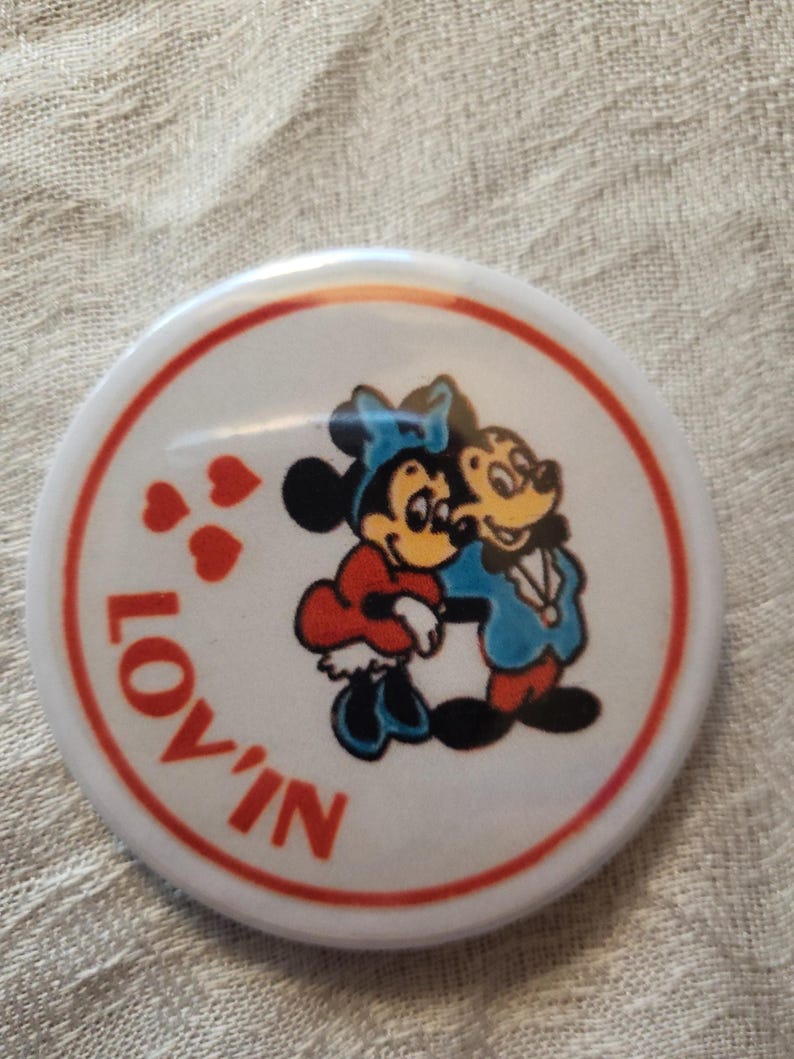 Op de afbeelding: Een ronde witte knoop met een rode rand en het woord "LOV'IN" in het rood. De knoop toont een cartoonafbeelding van Mickey en Minnie Mouse die elkaar omhelzen, met rode harten. De personages dragen blauwe, rode en gele kleding.