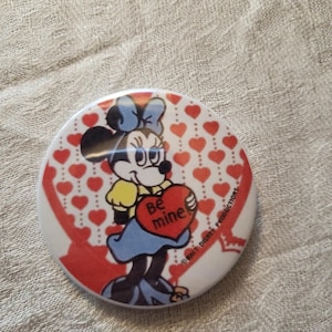 Op de afbeelding: Een ronde knoop met Minnie Mouse die een hart vasthoudt met de tekst "Be mine". De knoop heeft een witte achtergrond met rode harten en een rood lintontwerp. Een Disney-product.