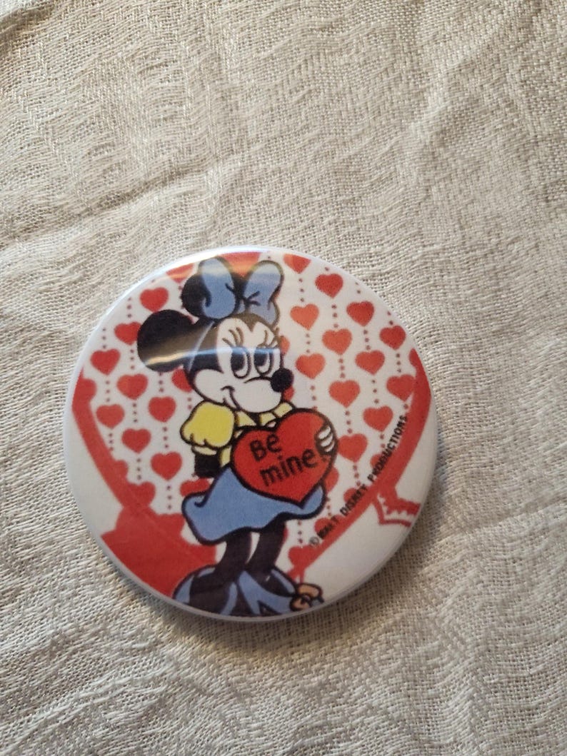 Op de afbeelding: Een ronde knoop met Minnie Mouse die een hart vasthoudt met de tekst "Be mine". De knoop heeft een witte achtergrond met rode harten en een rood lintontwerp. Een Disney-product.