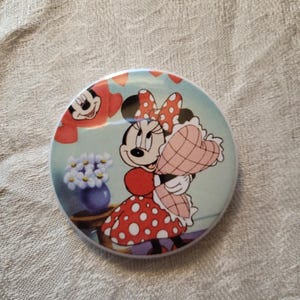 Op de afbeelding: Een ronde knoop met een illustratie van Minnie Mouse in een rode jurk met stippen en strik, met een hartvormig cadeau. De achtergrond is lichtblauw met een vaas met bloemen en Mickey Mouse. De knoop heeft een glanzende afwerking.