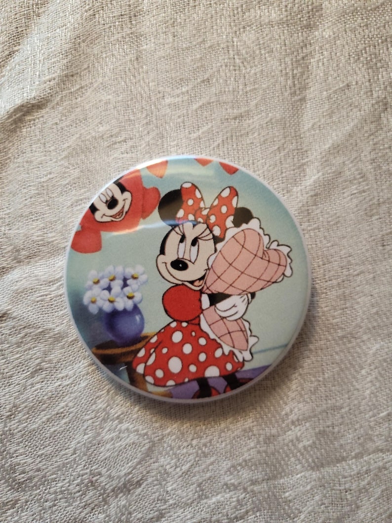 Op de afbeelding: Een ronde knoop met een illustratie van Minnie Mouse in een rode jurk met stippen en strik, met een hartvormig cadeau. De achtergrond is lichtblauw met een vaas met bloemen en Mickey Mouse. De knoop heeft een glanzende afwerking.