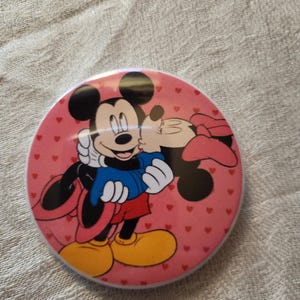 Op de afbeelding: Een ronde, roze button met Mickey en Minnie Mouse in een liefdevolle omhelzing. Mickey draagt een blauwe jas, rode shorts en gele schoenen. Minnie draagt een roze strik. De achtergrond is bedekt met kleine rode hartjes.