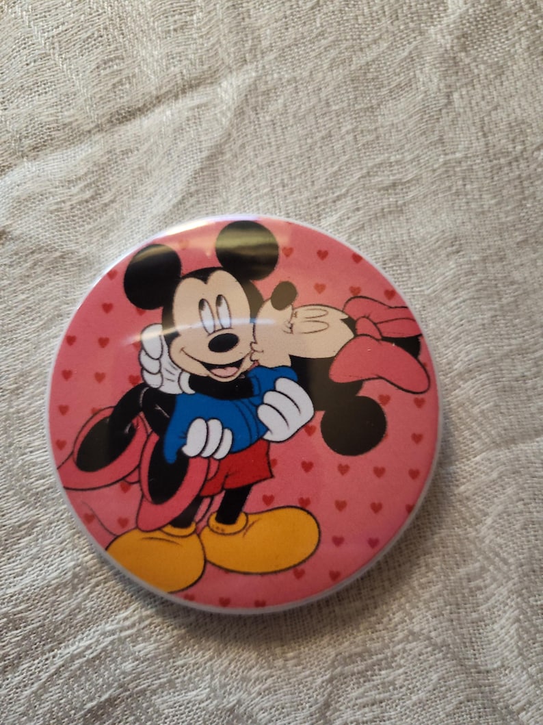 Op de afbeelding: Een ronde, roze button met Mickey en Minnie Mouse in een liefdevolle omhelzing. Mickey draagt een blauwe jas, rode shorts en gele schoenen. Minnie draagt een roze strik. De achtergrond is bedekt met kleine rode hartjes.