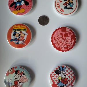 Op de afbeelding: Zes ronde Disney-personagebuttons met Mickey en Minnie Mouse. De buttons hebben Valentijnsdag- en hartthema's, met tekst als "Lov'in" en "Be mine".