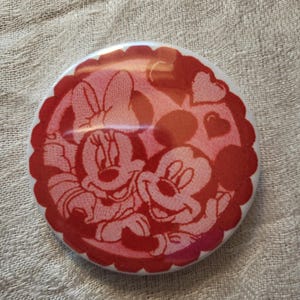 Op de afbeelding: Een ronde, rood-witte knoop met Mickey en Minnie Mouse die elkaar omarmen. De knoop heeft een geschulpte rand en een hartontwerp op de achtergrond. De afbeelding is in cartoonstijl.