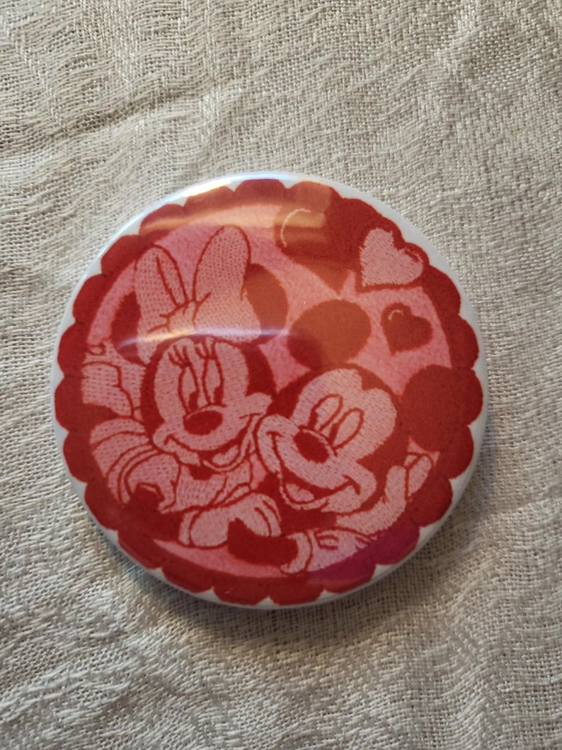 Op de afbeelding: Een ronde, rood-witte knoop met Mickey en Minnie Mouse die elkaar omarmen. De knoop heeft een geschulpte rand en een hartontwerp op de achtergrond. De afbeelding is in cartoonstijl.