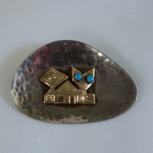 May include: A silver and gold brooch with a stylized cat design. The cat's eyes are turquoise blue.