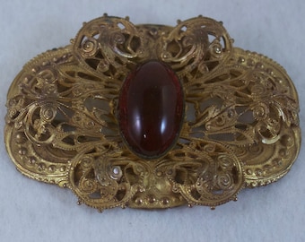 Op de afbeelding: Een gouden broche met een rode glazen cabochon in een filigraanontwerp.