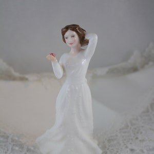 May include: A porcelain figurine of a woman in a long white dress, holding a small pink object. The woman has brown hair and is posed with one hand touching her hair. The figurine is set on a white surface with lace trim.
