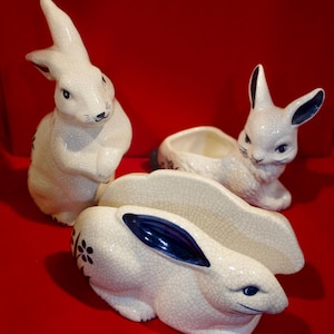 May include: Three white ceramic bunny figurines with blue accents. The bunnies are in a variety of poses, including sitting, standing, and lying down. The figurines are all made of a crackled glaze.