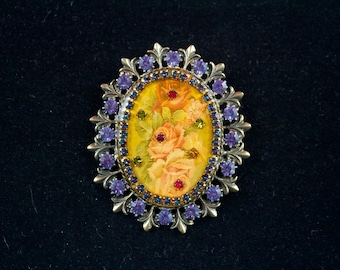 Può includere: Una spilla ovale con un pannello centrale giallo caratterizzato da un disegno floreale dipinto di rose arancioni. Il centro è circondato da un bordo di piccoli strass blu, accentuato da pietre viola più grandi e una cornice in filigrana.