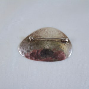 May include: Silver brooch with a hammered finish and a rounded shape. The back of the brooch has a maker's mark that reads "925 Sterling 12/14 TM".
