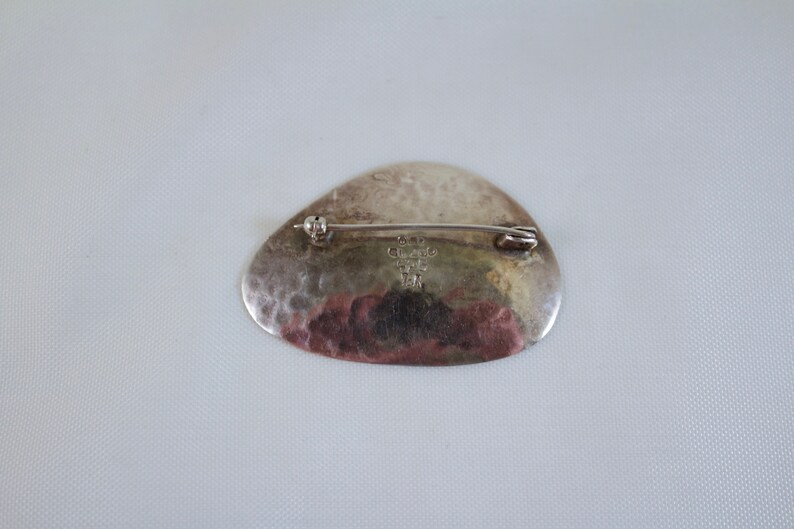 May include: Silver brooch with a hammered finish and a rounded shape. The back of the brooch has a maker's mark that reads "925 Sterling 12/14 TM".