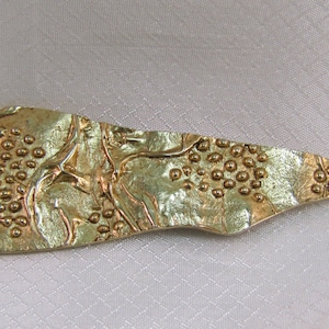 May include: A gold-toned metal pendant with a textured surface and a raised design. The pendant is shaped like a long, thin leaf with a rounded tip.