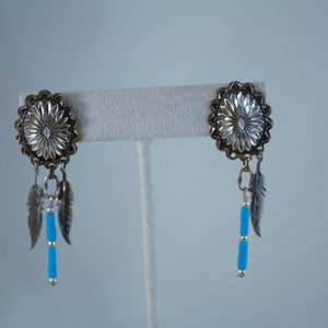 May include: A pair of silver earrings with turquoise beads and feather charms. The earrings are round and have a floral design.