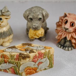 May include: A collection of five small animal figurines and one rectangular box. The figurines include a sheep, a cat with a bow, a gray puppy, a lion, and a pug holding a gift with the text Happy Birthday. The box features a relief of dolphins.