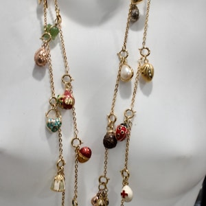May include: A multi-strand gold-tone necklace featuring a variety of colourful charms. The charms include spheres, teardrops, and bell shapes in red, white, green, and gold. The necklace is displayed on a mannequin, showcasing its layered design.