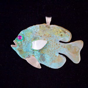 May include: A turquoise blue and green fish pendant with a silver eye and a white shell detail. The pendant is made of metal and has a textured surface.
