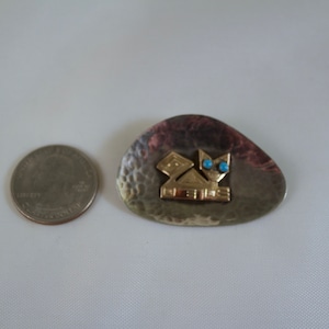 May include: A silver and gold brooch with a geometric design and turquoise accents. The brooch is shaped like a teardrop and has a hammered texture. The text "NEVIS" is inscribed on the brooch.