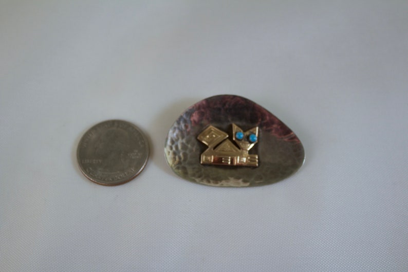 May include: A silver and gold brooch with a geometric design and turquoise accents. The brooch is shaped like a teardrop and has a hammered texture. The text "NEVIS" is inscribed on the brooch.