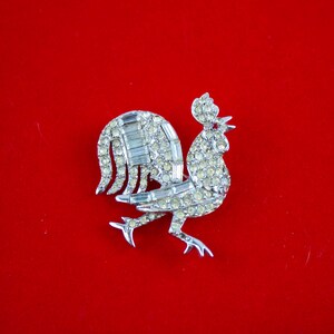 May include: A silver-toned rhinestone rooster brooch. The rooster is facing right and has a detailed design with clear rhinestones.