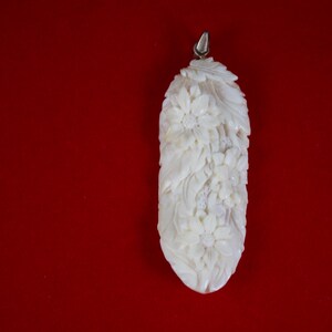 May include: Ivory-colored pendant with a floral design. The pendant is elongated with a silver-colored bail at the top. The carved flowers and leaves create a textured surface against a red background.