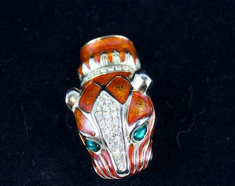 Puede incluir: Broche esmaltado detallado con forma de cabeza estilizada de felino. El broche presenta tonos naranja, rojo y plateado, con cristales transparentes brillantes y ojos verde esmeralda. La parte superior tiene un diseño de corona.