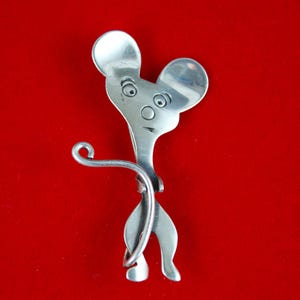 Puede incluir: Una escultura metálica caprichosa con forma de ratón, hecha de cubiertos reciclados. La escultura presenta cabezas de cuchara para las orejas y un mango de tenedor para el cuerpo. La pieza tiene un acabado plateado pulido y está sobre un fondo rojo.