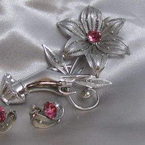 May include: A silver filigree brooch featuring a hand holding a flower with a pink rhinestone centre. Matching earrings with pink rhinestones are also included.