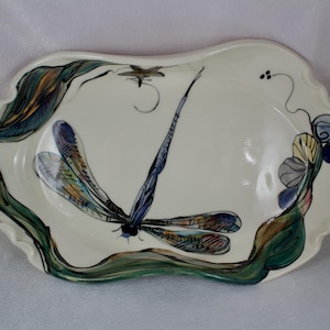 May include: A white ceramic serving tray with a hand-painted design of a dragonfly with blue, green, and yellow accents. The tray has a wavy edge and a leafy design around the border.