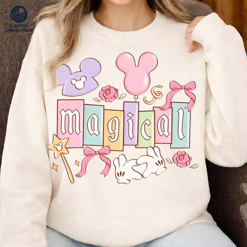 A woman wearing a white sweater with a colorful and whimsical design featuring the word "magical" surrounded by various Disney-themed elements such as Mickey Mouse ears, a balloon, and flowers.