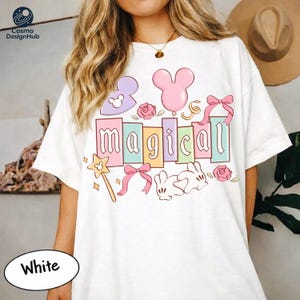 A woman wearing a white t-shirt with a colorful and whimsical design featuring the word "Magical" surrounded by various Disney-themed elements such as Mickey Mouse, balloons, and flowers.