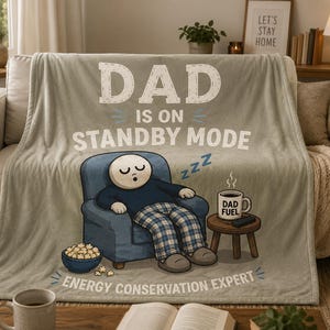 May include: A light beige fleece blanket featuring a cartoon illustration of a person sleeping in a blue armchair. The text reads DAD IS ON STANDBY MODE and ENERGY CONSERVATION EXPERT. A mug labeled DAD FUEL sits on a small wooden side table nearby.