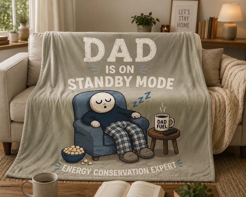 May include: A light beige fleece blanket featuring a cartoon illustration of a person sleeping in a blue armchair. The text reads DAD IS ON STANDBY MODE and ENERGY CONSERVATION EXPERT. A mug labeled DAD FUEL sits on a small wooden side table nearby.