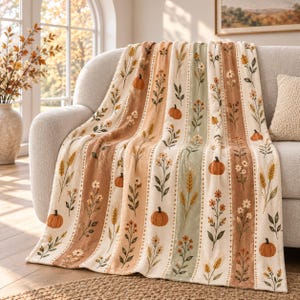 May include: A soft throw blanket featuring a vertical striped pattern with alternating bands of cream, tan, and sage green. The design includes illustrations of orange pumpkins, wheat stalks, and various wildflowers in shades of brown and green.