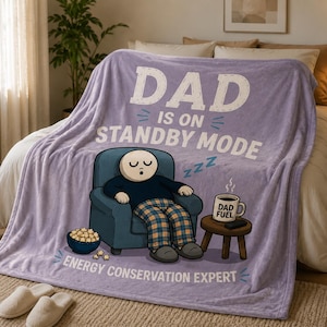 May include: A light purple fleece blanket featuring a cartoon illustration of a person sleeping in a blue armchair. The text reads DAD IS ON STANDBY MODE and ENERGY CONSERVATION EXPERT. A mug labeled DAD FUEL sits on a small wooden side table.