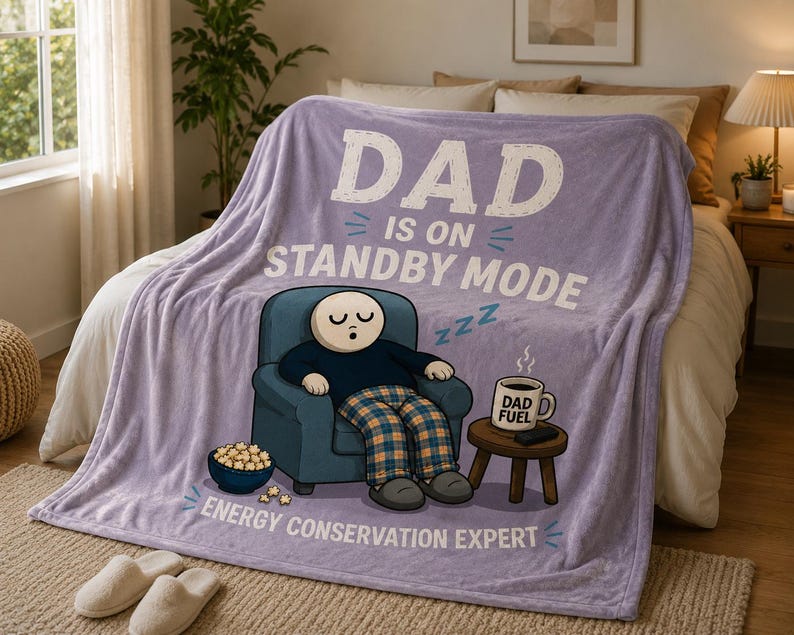 May include: A light purple fleece blanket featuring a cartoon illustration of a person sleeping in a blue armchair. The text reads DAD IS ON STANDBY MODE and ENERGY CONSERVATION EXPERT. A mug labeled DAD FUEL sits on a small wooden side table.
