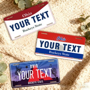 May include: Three Ohio license plates with the text "YOUR TEXT" in bold, white letters. The plates have different designs, including red and white, blue and red, and a music-themed design with a guitar and microphone. The words "Ohio" and "Buckeye State" are also visible.