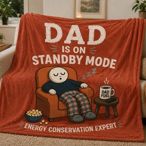 May include: A rust-colored fleece throw blanket featuring the text DAD IS ON STANDBY MODE and ENERGY CONSERVATION EXPERT. The graphic shows a character sleeping in an orange armchair next to a bowl of popcorn and a mug labeled DAD FUEL.