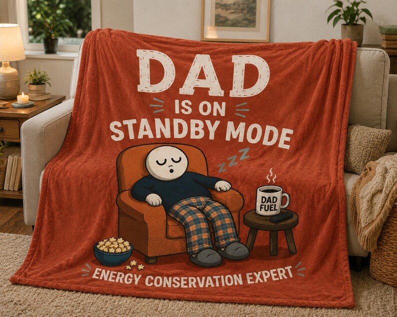 May include: A rust-colored fleece throw blanket featuring the text DAD IS ON STANDBY MODE and ENERGY CONSERVATION EXPERT. The graphic shows a character sleeping in an orange armchair next to a bowl of popcorn and a mug labeled DAD FUEL.