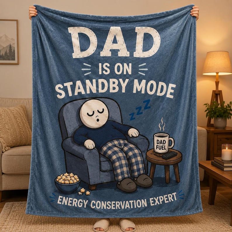 May include: A blue fleece blanket featuring the white text DAD IS ON STANDBY MODE and ENERGY CONSERVATION EXPERT. The graphic shows a cartoon character sleeping in a blue armchair next to a small table with a mug labeled DAD FUEL and a bowl of popcorn.