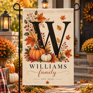 May include: A rectangular garden flag featuring a large black letter W, an orange pumpkin, a white pumpkin, and autumn leaves. The text reads WILLIAMS family and EST. 2020. The design is printed on a cream-colored fabric background.