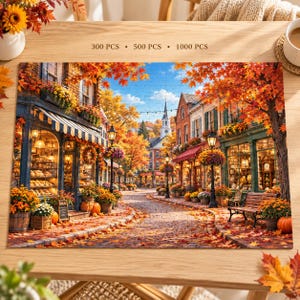 May include: A jigsaw puzzle depicting a cobblestone street scene in autumn with shops, orange and yellow trees, a church steeple in the distance, and pumpkins. The text 300 PCS, 500 PCS, and 1000 PCS is printed above the puzzle on the wooden table.