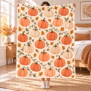 May include: A cream-colored throw blanket featuring a repeating pattern of orange and white pumpkins with green vines and scattered autumn leaves. The design is printed across the entire surface of the rectangular fabric, showcasing various pumpkin sizes.