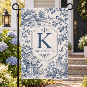 May include: A rectangular garden flag featuring a blue and white toile pattern with a central monogram letter K and the text THE KENNEDYS. The design includes a barn, a bird on a branch, butterflies, a rabbit, and a metal watering can.